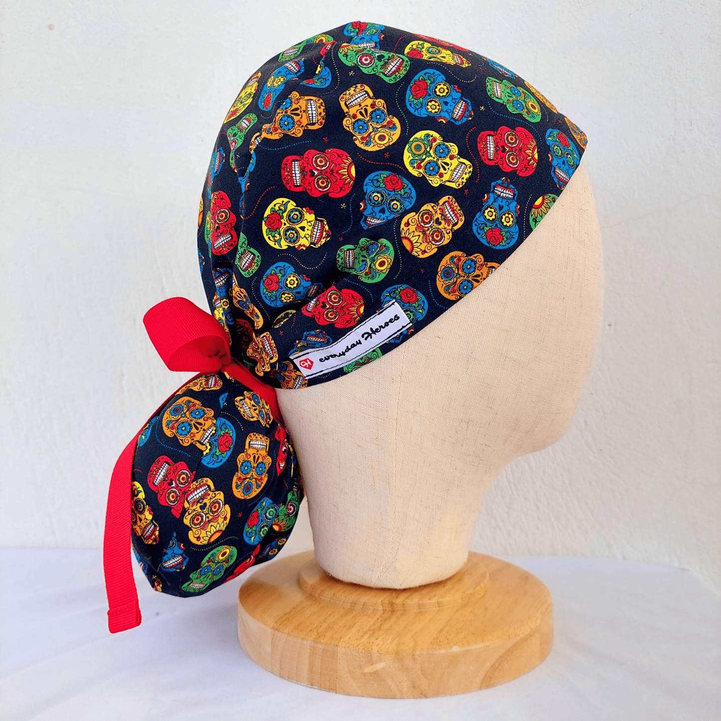 Left side of a mannequin wearing a scrub cap ponytail with small colorful skulls and a separate pounch on it for the ponytail that hold the long hair and ties with red ribbons