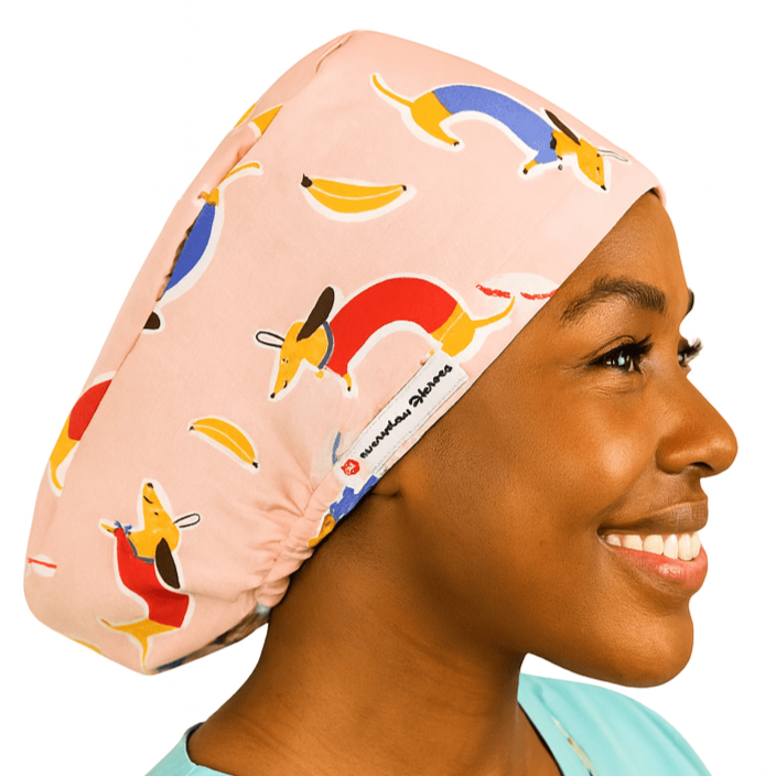 Custom Doxie dachshund vet scrub cap for women, shown on a mannequin head, featuring a playful weenie dog and sausage dog print in pink tones. This euro-style scrub cap is designed for animal surgeons and veterinary professionals. Optional satin lining adds comfort, with additional customization available including buttons for mask support and personalized embroidery. A fun and functional surgical scrub hat with ponytail space.