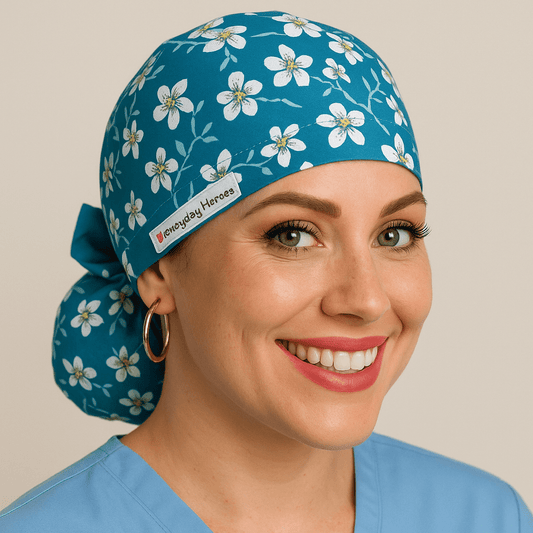 Almond floral women’s scrub cap with white flower print on teal fabric, ponytail style with bow tie back and Everyday Heroes label. Satin lined medical surgical hat, modeled by a smiling nurse in a light blue scrub top with gold hoop earrings. Personalized scrub cap, reusable and washable, perfect gift for doctors, nurses, or as a graduation gift for healthcare professionals.