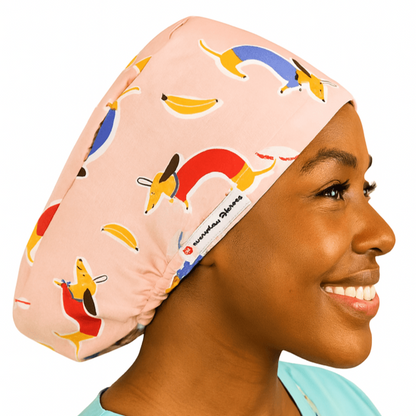 Custom Doxie dachshund vet scrub cap for women, shown on a mannequin head, featuring a playful weenie dog and sausage dog print in pink tones. This euro-style scrub cap is designed for animal surgeons and veterinary professionals. Optional satin lining adds comfort, with additional customization available including buttons for mask support and personalized embroidery. A fun and functional surgical scrub hat with ponytail space.