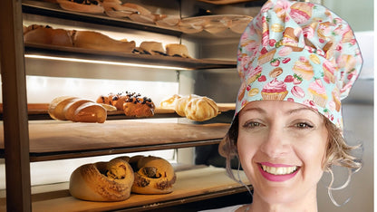 a smilling woman wearing the chefs hat withcupcakes as she working on abakery.